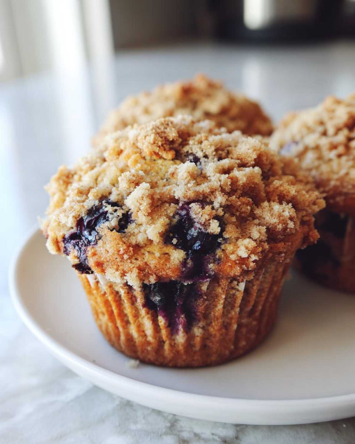 A close-up of a fresh blueberry muffin with a golden crumb topping sitting on a white plate.