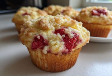 A close-up of a freshly baked strawberry muffin topped with sweet crumble, with more strawberry muffins visible in the background.