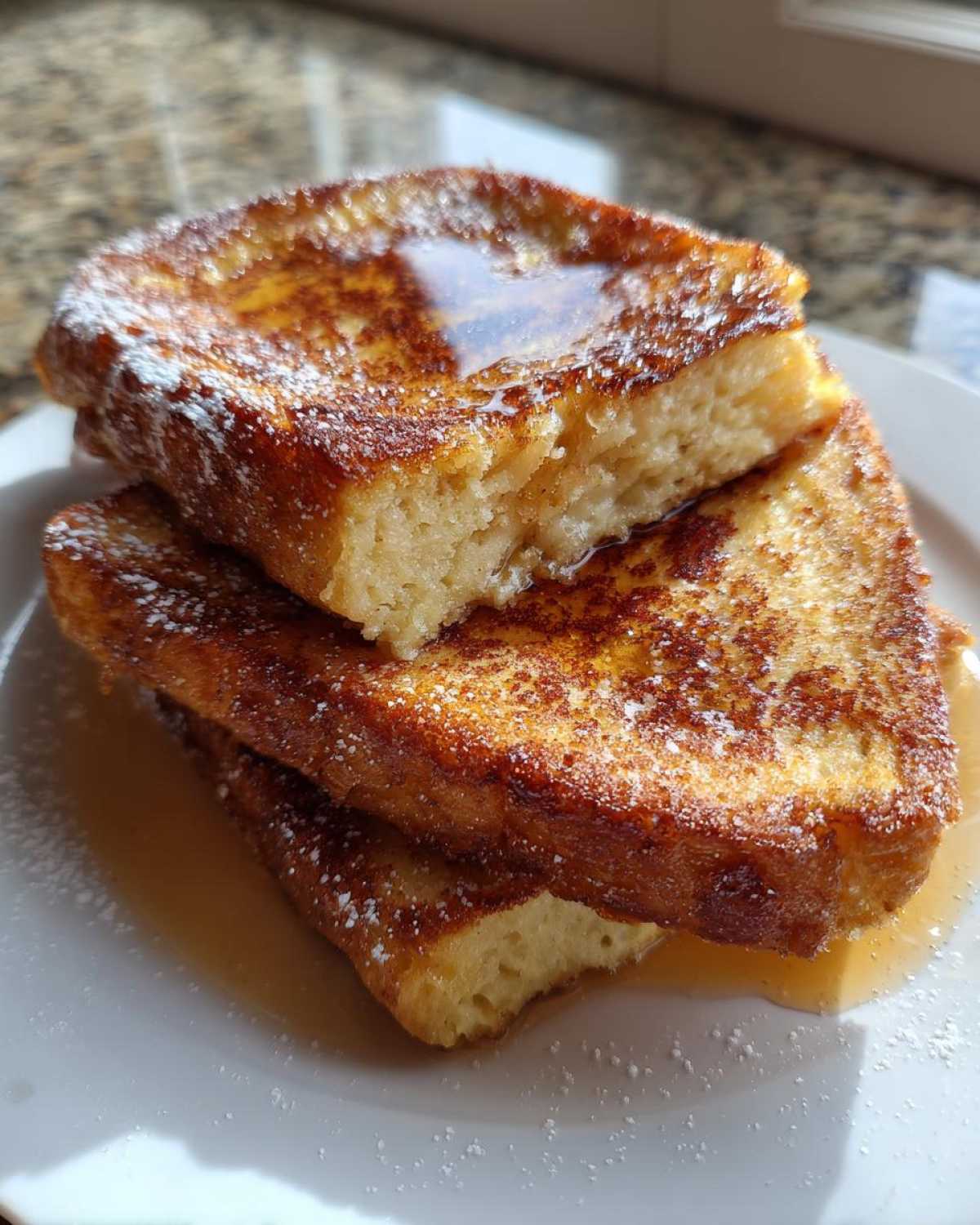 A close-up of three stacked slices of golden brown easy french toast drizzled with syrup and dusted with powdered sugar.