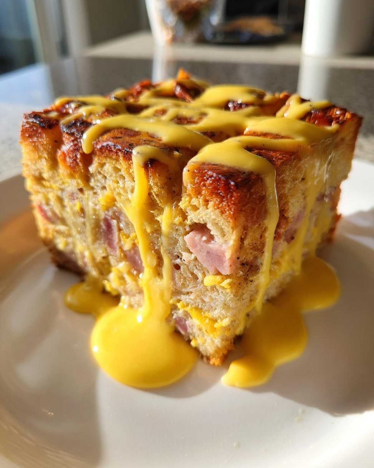 A thick slice of eggs benedict casserole featuring bread, ham, and cheese, generously drizzled with bright yellow hollandaise sauce.