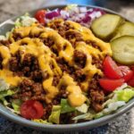 A close-up view of a ready-to-eat cheeseburger bowl featuring seasoned ground beef, cheese sauce, lettuce, pickles, and tomatoes.