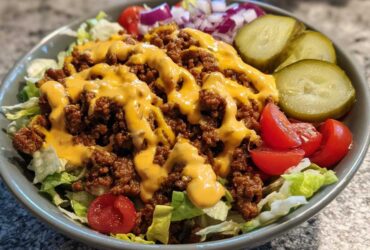 A close-up view of a ready-to-eat cheeseburger bowl featuring seasoned ground beef, cheese sauce, lettuce, pickles, and tomatoes.