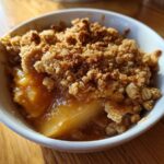 A close-up view of a warm serving of pear crisp in a white bowl, featuring bubbly fruit filling and a crunchy oat topping.