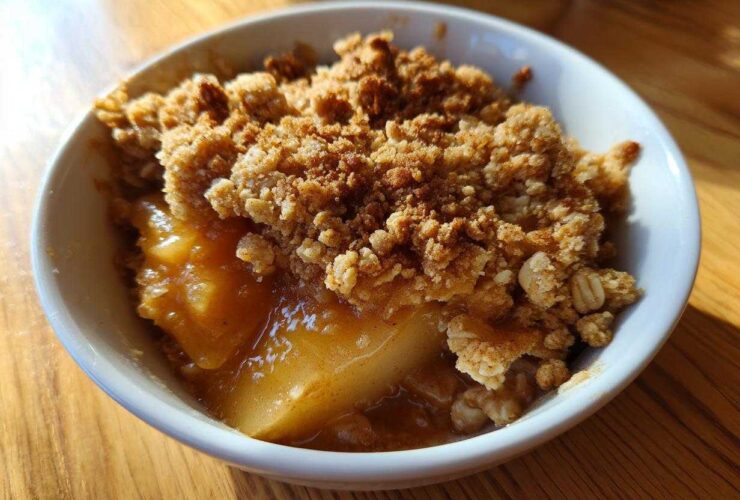 A close-up view of a warm serving of pear crisp in a white bowl, featuring bubbly fruit filling and a crunchy oat topping.