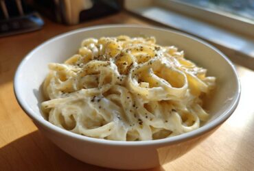 A white bowl filled with fettuccine pasta generously coated in creamy homemade alfredo sauce and sprinkled with herbs.