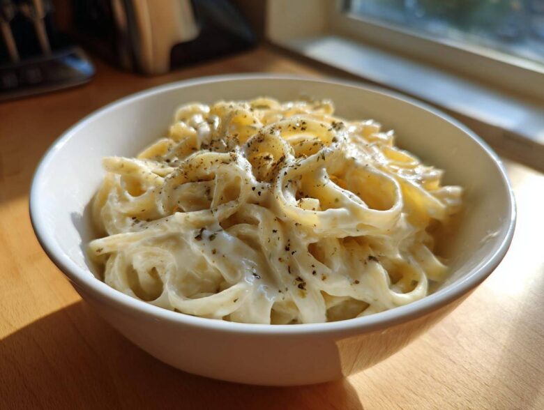 A white bowl filled with fettuccine pasta generously coated in creamy homemade alfredo sauce and sprinkled with herbs.