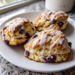 Three freshly baked blueberry scones topped with a sweet vanilla glaze, served on a white plate.