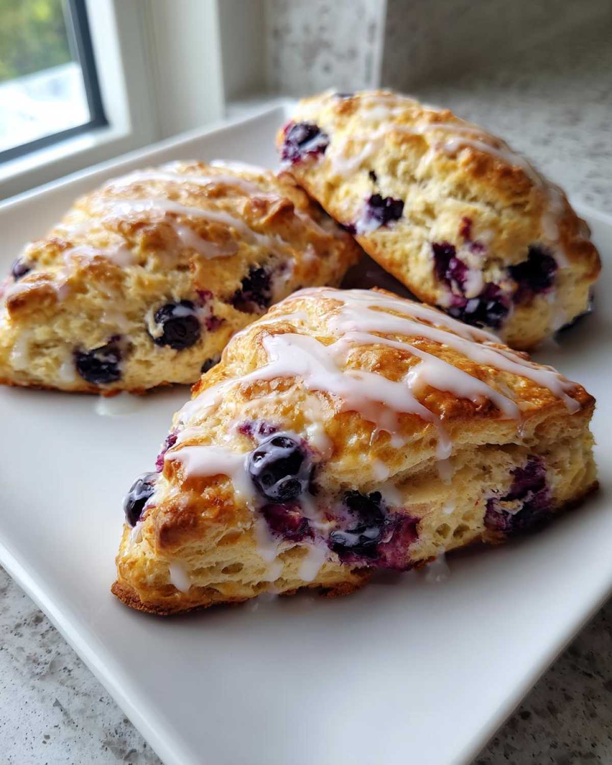 Three flaky blueberry scones drizzled with white vanilla glaze, resting on a white rectangular plate.