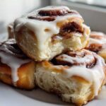 A stack of freshly baked, fluffy cinnamon roll pastries generously topped with dripping white cream cheese icing.