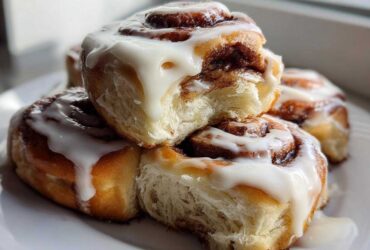 A stack of freshly baked, fluffy cinnamon roll pastries generously topped with dripping white cream cheese icing.