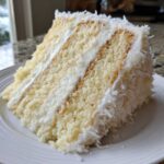 Close-up of a moist slice of fluffy coconut cake layered with white frosting and covered in shredded coconut.