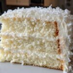 Close-up of a thick slice of fluffy coconut cake layered with white frosting and topped with shredded coconut.