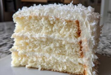 Close-up of a thick slice of fluffy coconut cake layered with white frosting and topped with shredded coconut.
