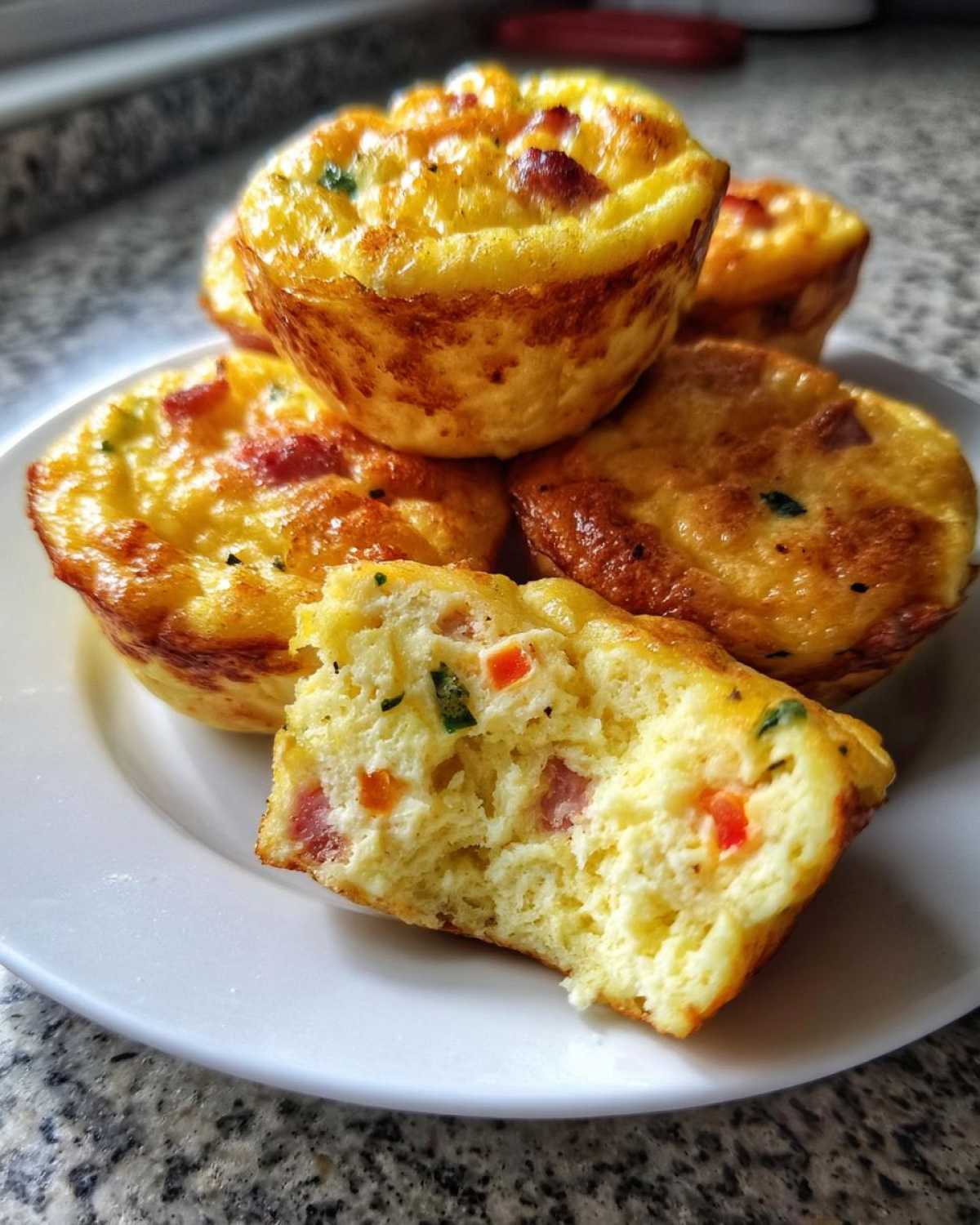 Close-up of fluffy muffin tin egg bites stacked on a white plate, one is broken open showing ham and peppers.