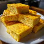 Close-up of stacked, fluffy, golden yellow portions of baked sheet pan eggs on a white plate.