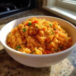 A close-up of a white bowl filled with fluffy, orange-colored spanish rice mixed with peas and diced red peppers.