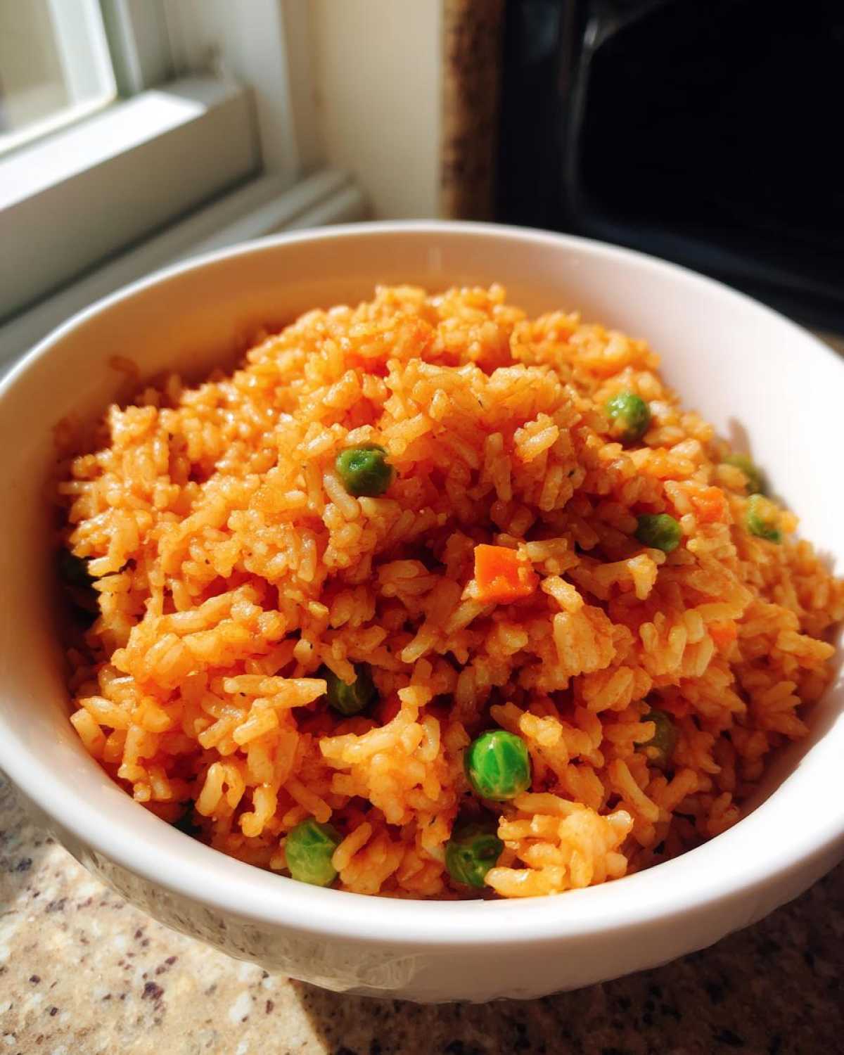 Close-up of a white bowl filled with fluffy, orange-colored spanish rice mixed with bright green peas and diced carrots.