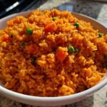 A close-up shot of a white bowl filled with fluffy, bright orange spanish rice mixed with green peas and diced carrots.