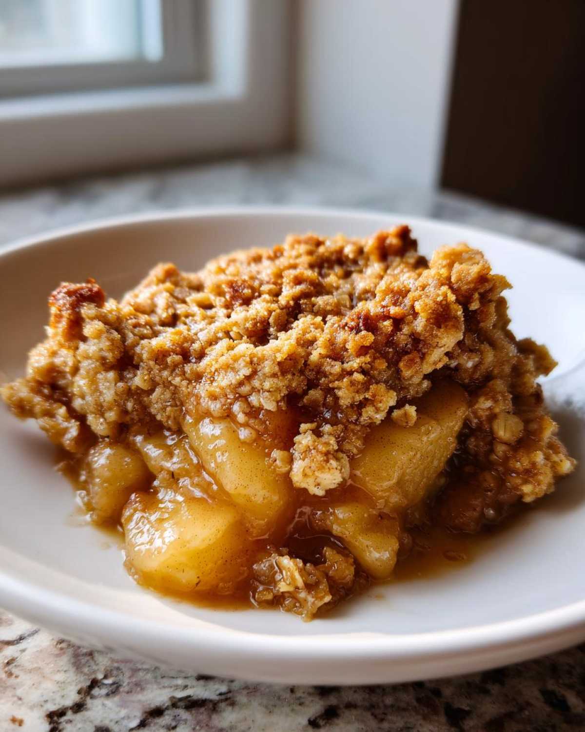 A close-up of a warm serving of apple crisp showing tender, saucy apples beneath a thick, golden-brown, crunchy topping.