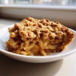 A close-up shot of a generous slice of apple crisp showing tender, spiced apples beneath a thick, golden-brown, crunchy topping.