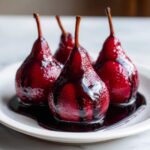 Four glistening, deep red wine poached pears served on a white plate with rich syrup.