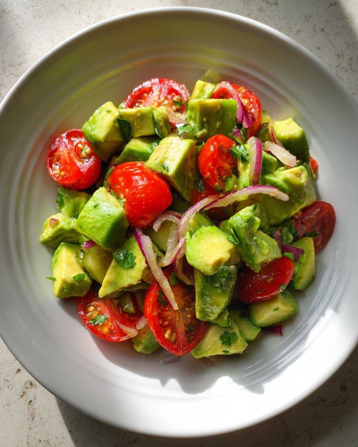 Close-up overhead view of a vibrant avocado salad with cherry tomatoes and red onion slices in a white bowl.