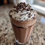 A tall glass filled with creamy frozen hot chocolate, topped with whipped cream and chocolate shavings.