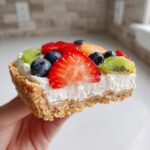 A hand holding a delicious slice of fruit pizza sugar cookie with fresh strawberries, blueberries, and kiwi.