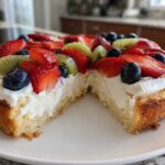 Close-up of a slice of fruit pizza sugar cookie showing the thick sugar cookie base, cream cheese frosting, strawberries, blueberries, and kiwi.