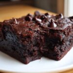 Close-up of a rich, fudgy slice of black bean brownies topped with chocolate chips on a white plate.