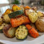 A close-up of perfectly cooked Garlic Herb Roasted Veggies, including potatoes, carrots, and zucchini, piled on a white plate.