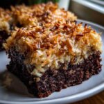 A moist slice of german chocolate poke cake featuring a rich chocolate base and thick, toasted coconut pecan frosting.