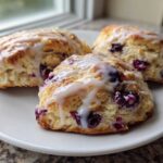 Three freshly baked blueberry scones topped with a sweet white glaze, resting on a white plate.