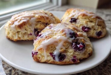 Three freshly baked blueberry scones topped with a sweet white glaze, resting on a white plate.