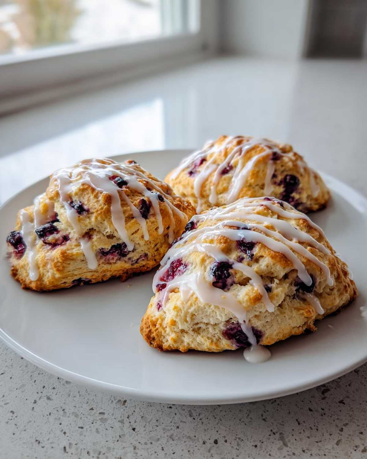 Three freshly baked blueberry scones, drizzled with white glaze, resting on a white plate.