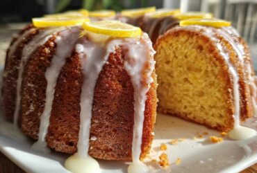 A glazed lemon pound cake, featuring a slice cut out to show the moist interior, topped with lemon slices.