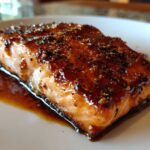 Close-up of a perfectly cooked, glazed salmon fillet resting in its sauce on a white plate.