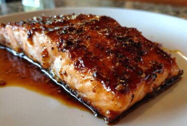 Close-up of a perfectly cooked, glazed salmon fillet resting in its sauce on a white plate.