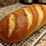 A freshly baked, golden brown loaf of french bread with deep slashes resting on a white wire cooling rack.