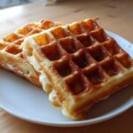 Two golden brown, freshly made high protein waffles stacked slightly on a white plate.