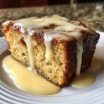 A close-up of a square slice of warm bread pudding topped generously with dripping vanilla custard sauce.