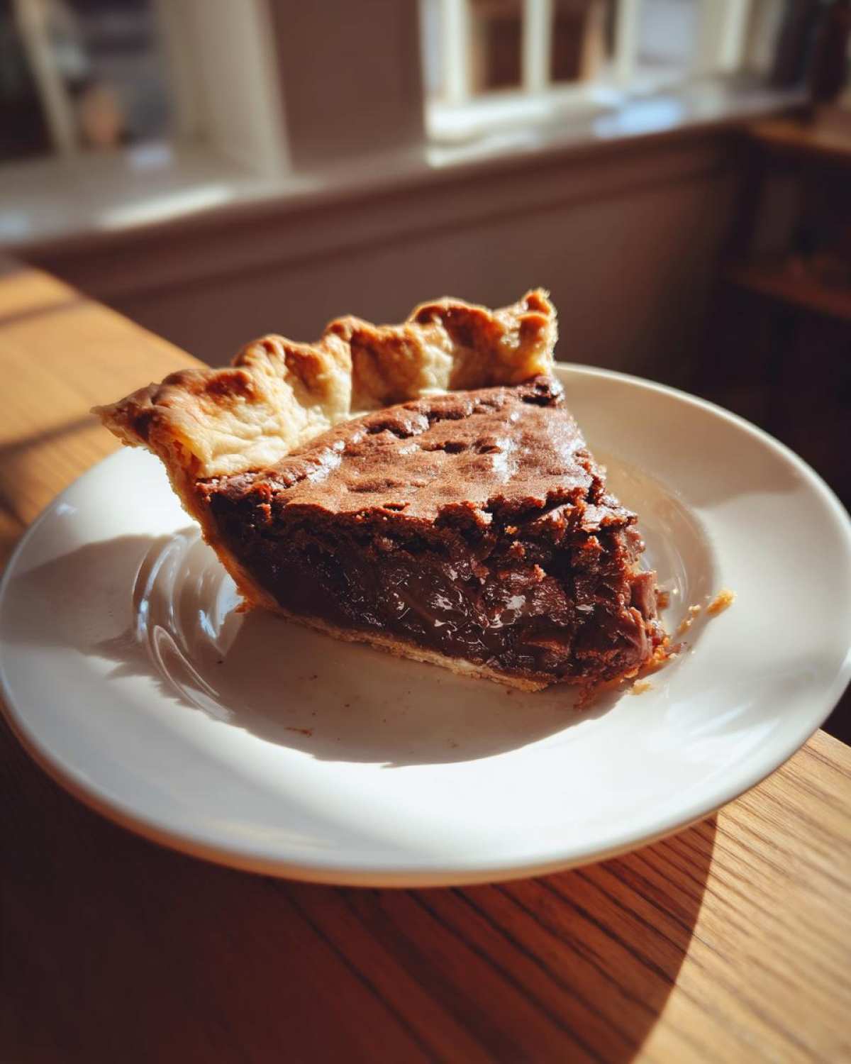 A close-up of a gooey slice of chocolate chess pie with a flaky crust, sitting on a white plate bathed in sunlight.