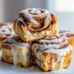 Close-up of three soft, fluffy cinnamon rolls stacked, dripping with thick white icing.