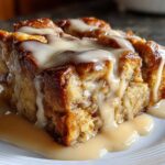 A close-up of a square slice of warm, gooey bread pudding topped generously with a vanilla glaze.