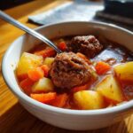 Close-up of a white bowl filled with rich, tomato-based albondigas soup featuring large meatballs, potatoes, and carrots.