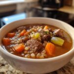 Close-up of a white bowl filled with rich beef barley soup, featuring chunks of beef, carrots, celery, and barley.