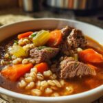 Close-up of a steaming bowl of rich beef barley soup featuring chunks of beef, carrots, and celery.