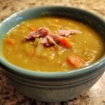 Close-up of a thick, yellow-green pea soup topped with shredded ham and carrots in a teal bowl.
