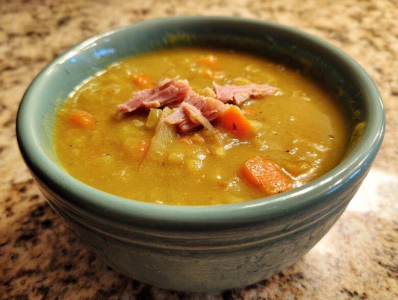 Close-up of a thick, yellow-green pea soup topped with shredded ham and carrots in a teal bowl.