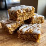 A stack of four square, dense oatmeal bars drizzled with white icing, sitting on a wooden cutting board.